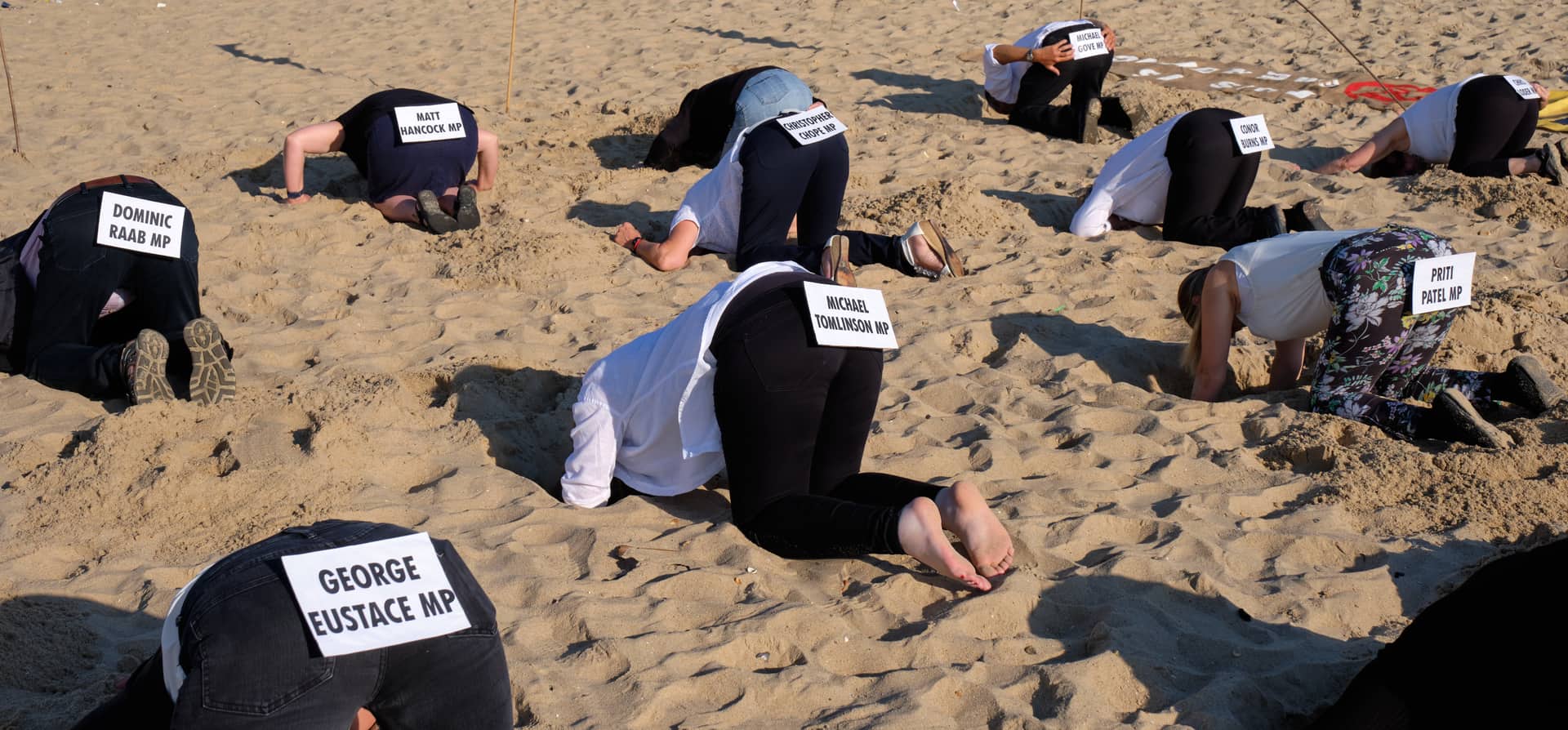 People kneeling on a beach with their heads buried in the sand, each with a card on their back naming an MP.